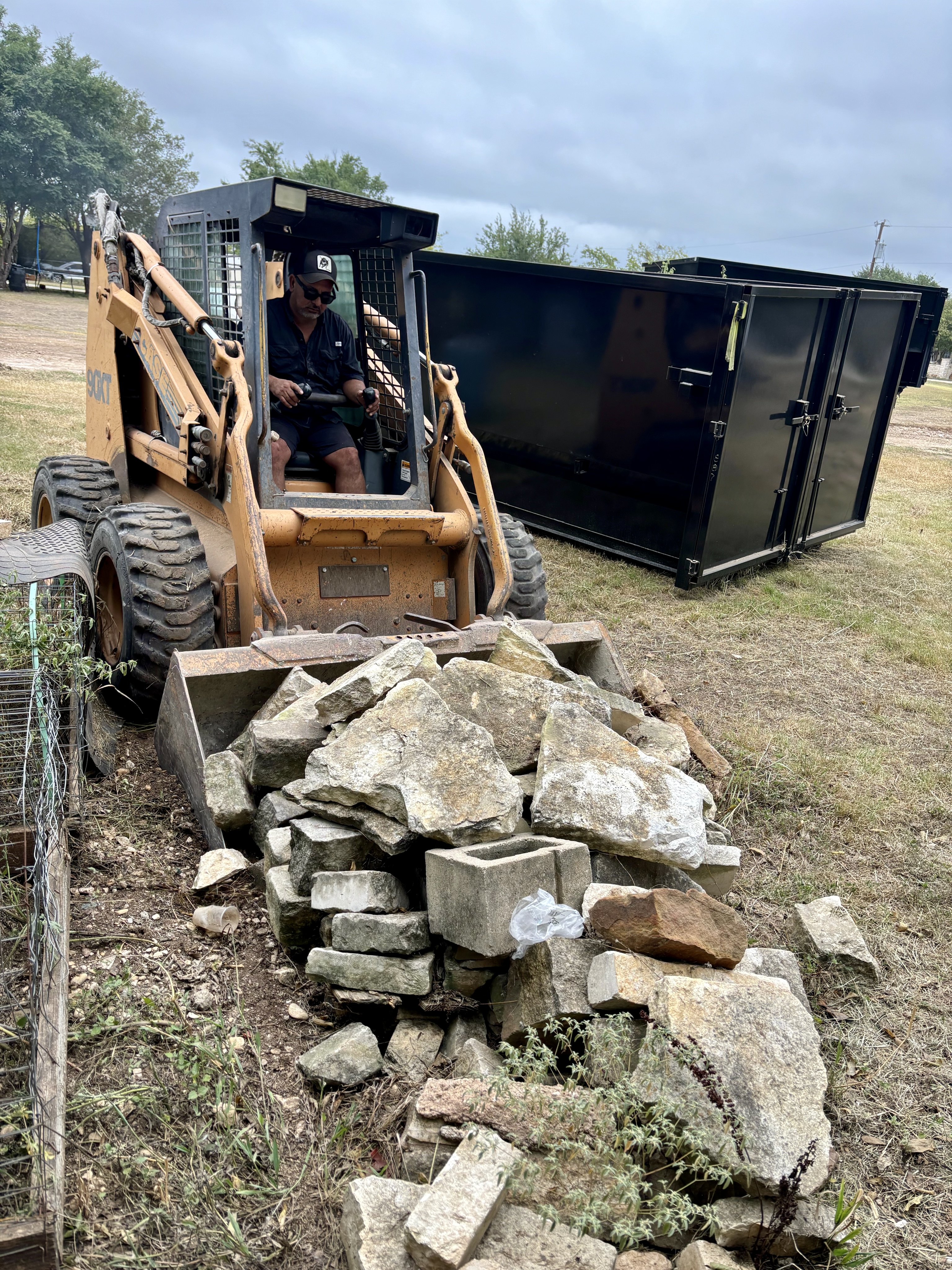V5 Inc. skid steer clearing rock debris at a job site with dumpster in background